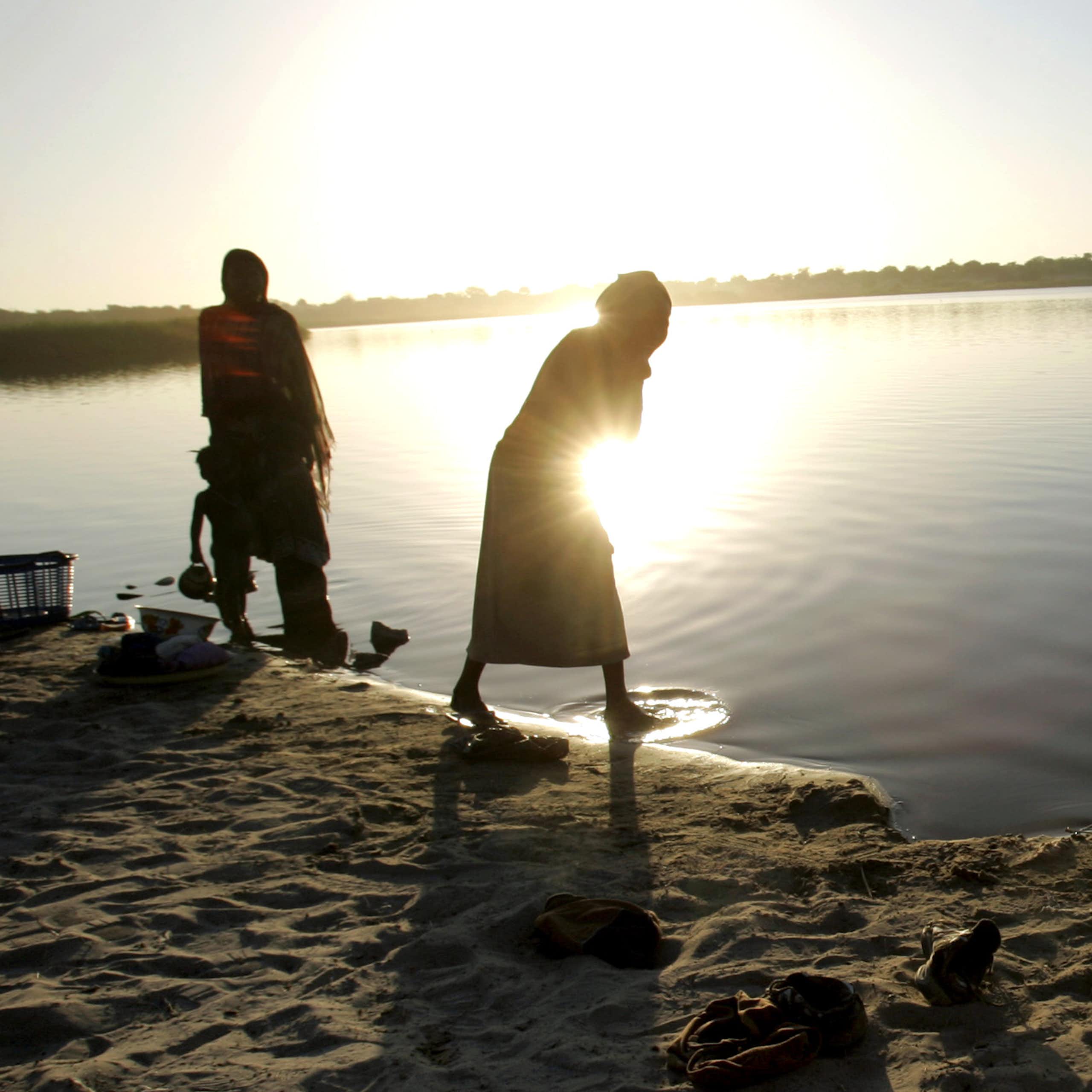 Pessoas lavam roupas na beira do Lago Chade com o pôr do sol sobre a água.