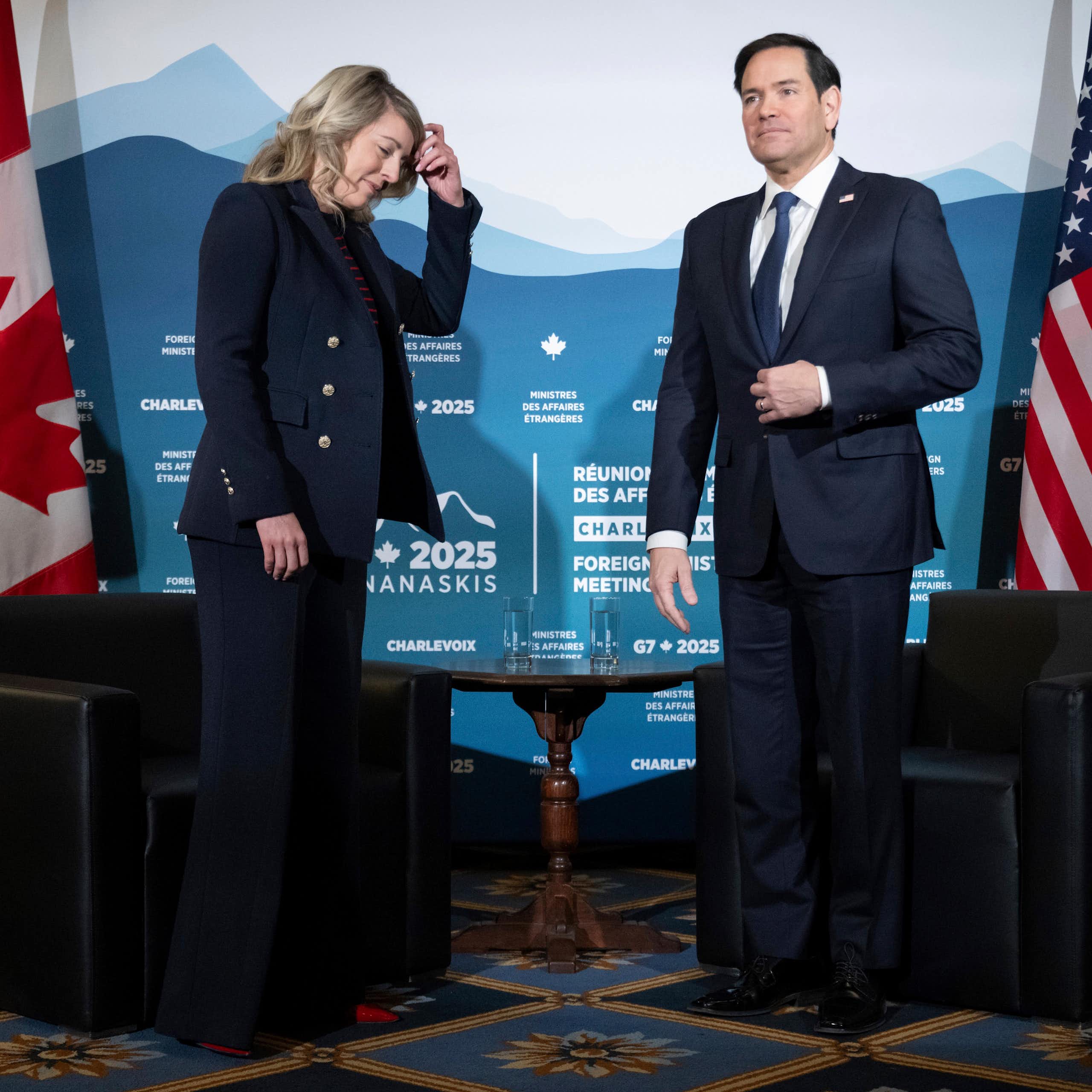 A woman with blonde shoulder-length hair has her hand on her head as she looks down. She's stands next to a dark-haired man. Canadian and U.S. flags are behind them.