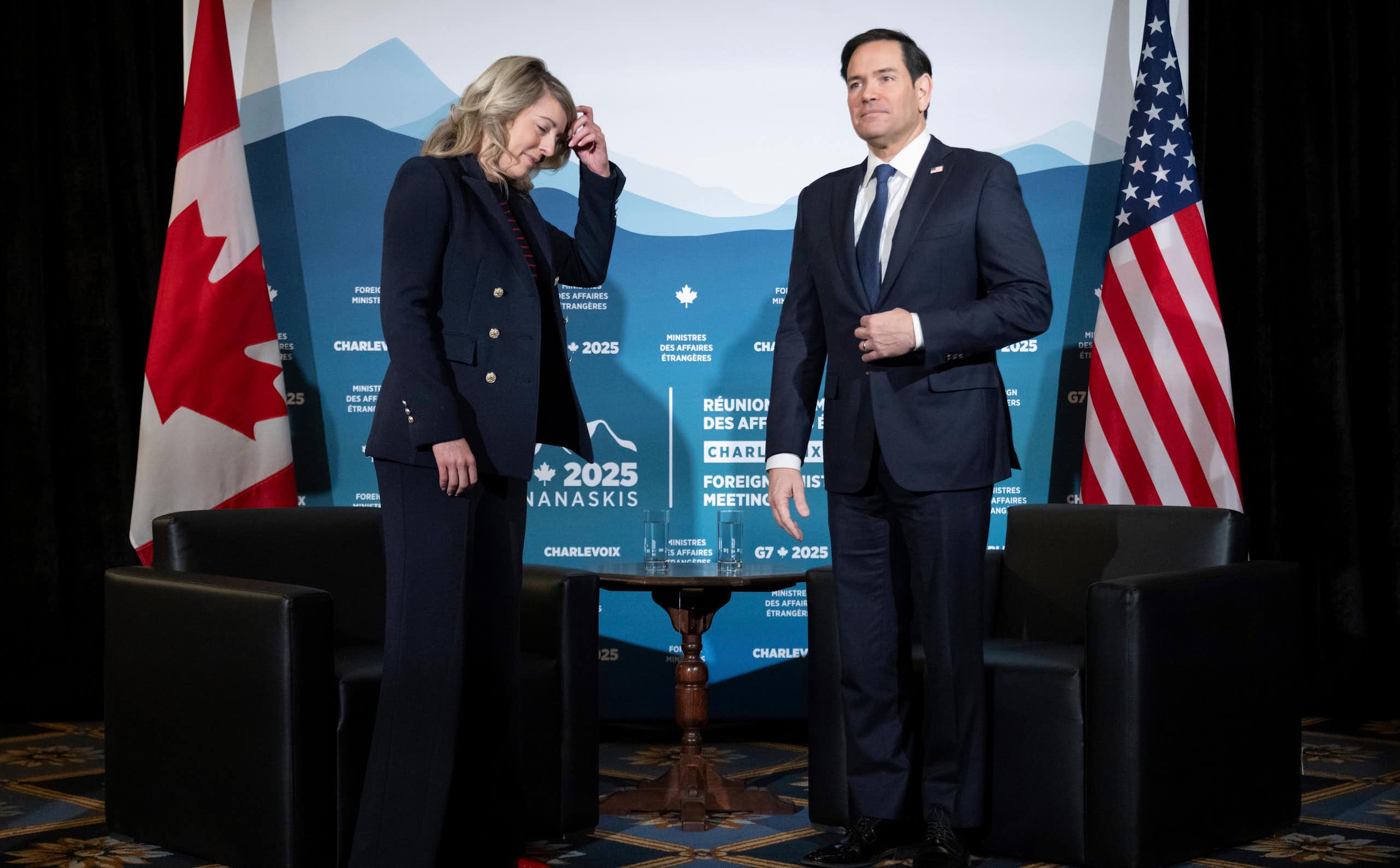 A woman with blonde shoulder-length hair has her hand on her head as she looks down. She's stands next to a dark-haired man. Canadian and U.S. flags are behind them.