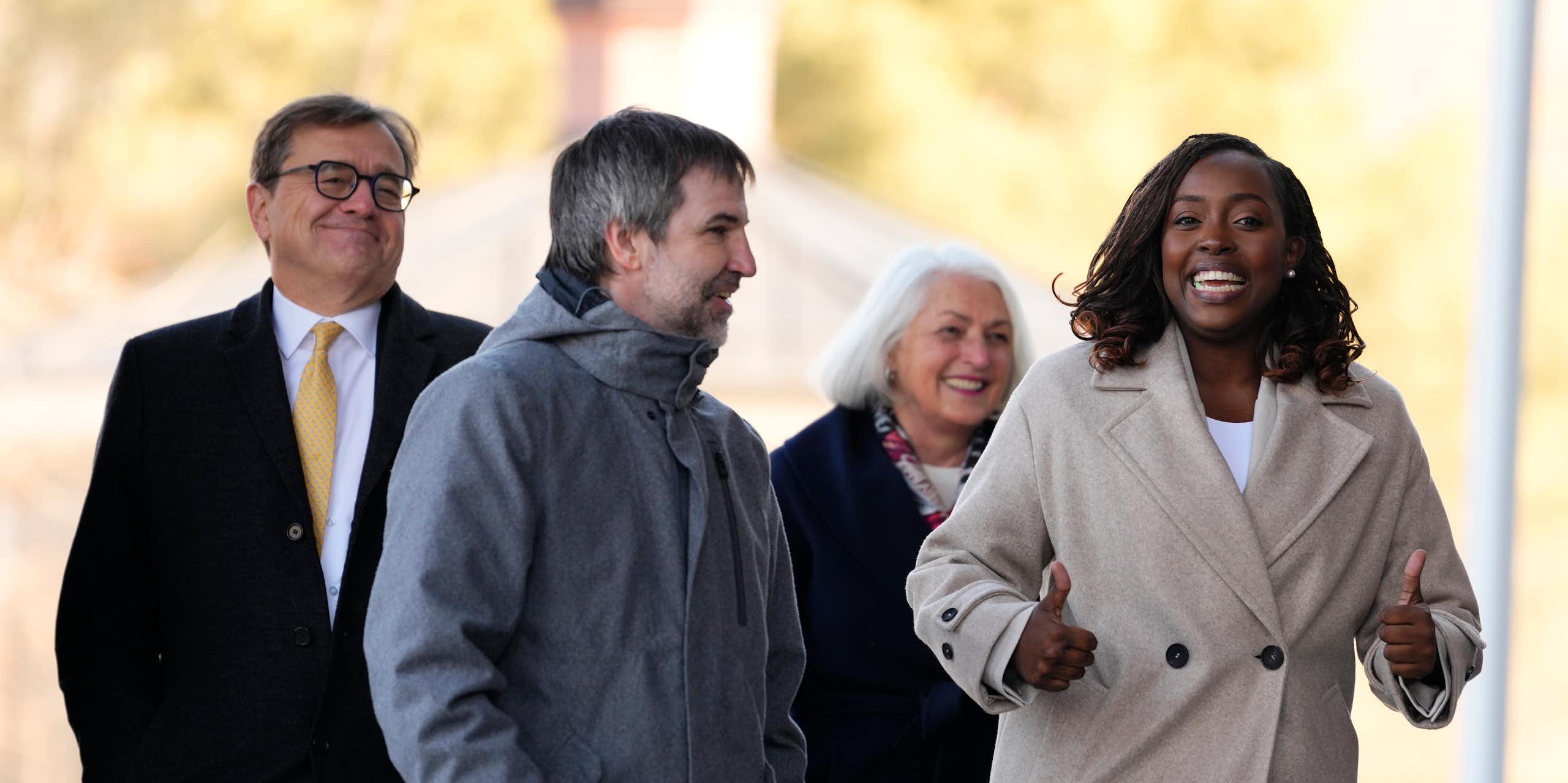 Two white men and a white woman walk with a Black woman. All are smiling and the Black woman is giving two thumbs up.