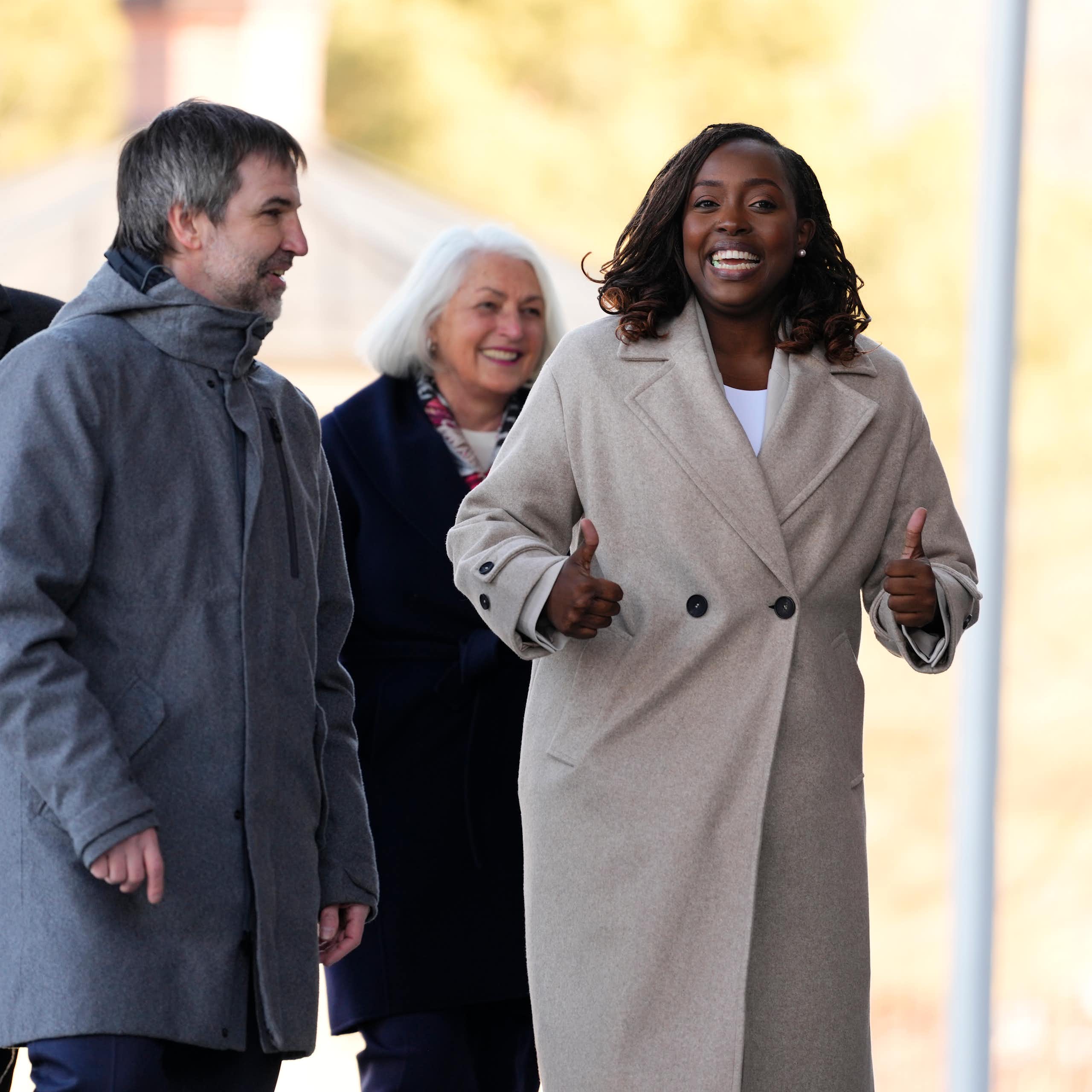 Two white men and a white woman walk with a Black woman. All are smiling and the Black woman is giving two thumbs up.