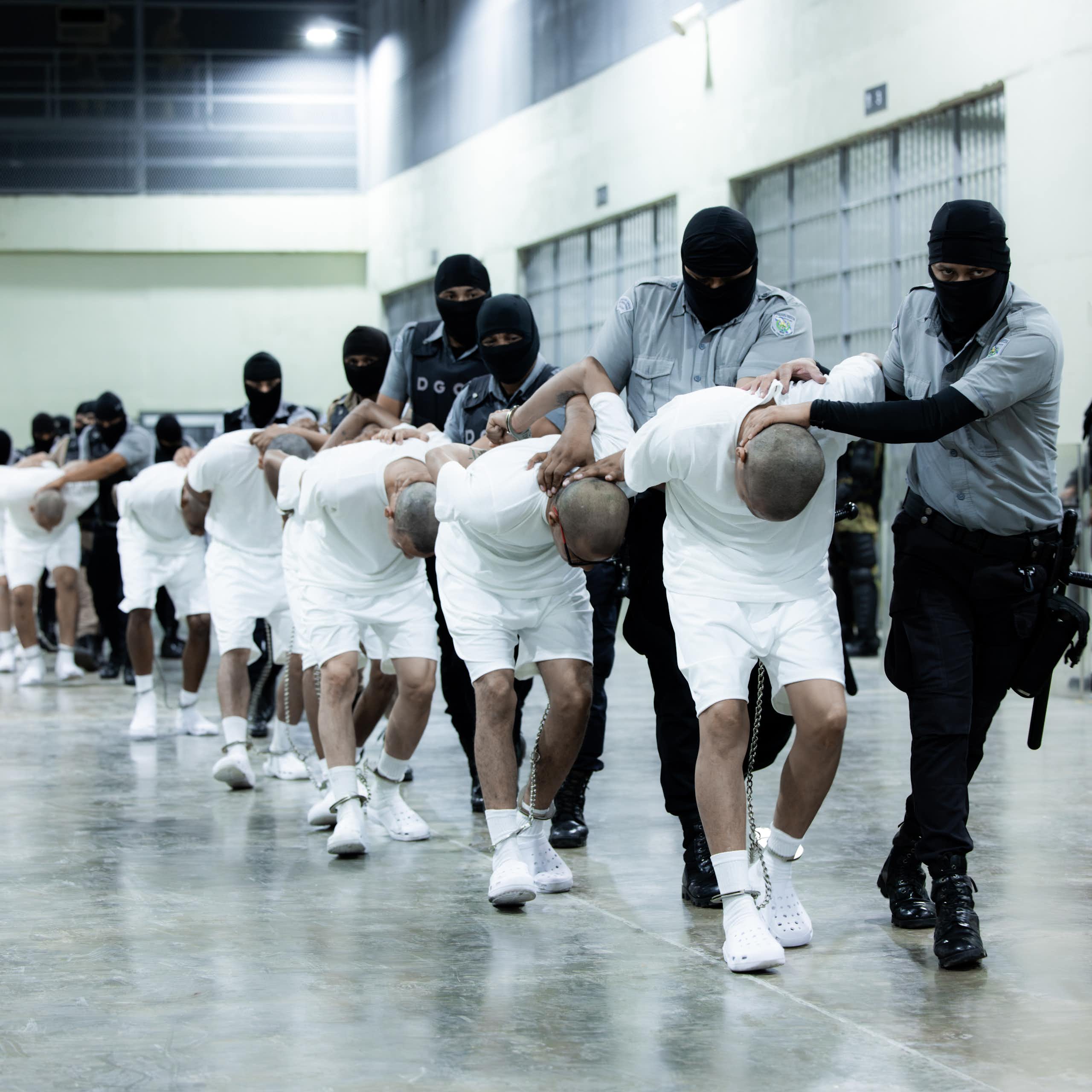 Men in white shirts and shorts are bent double, being led by arimed men in balaclavas