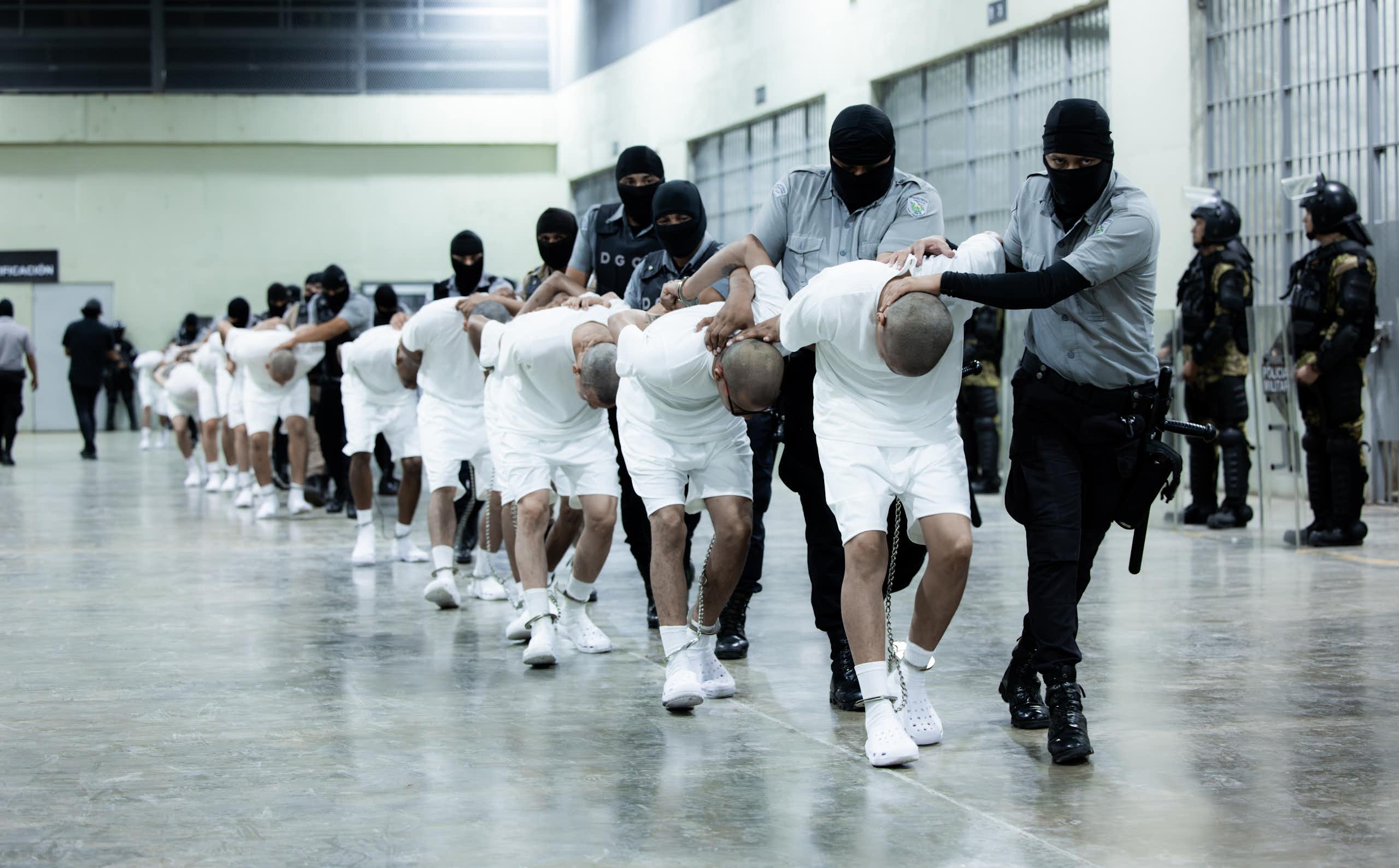Men in white shirts and shorts are bent double, being led by arimed men in balaclavas