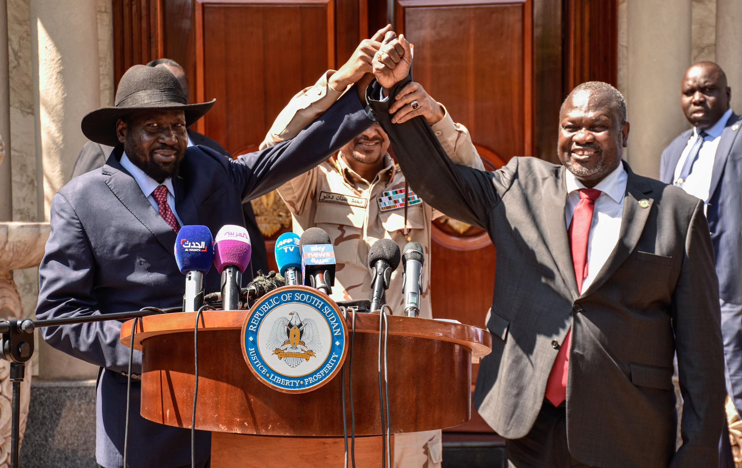 Two men have their hands held together in the middle by a third man as they stand behind a podium with microphones on it