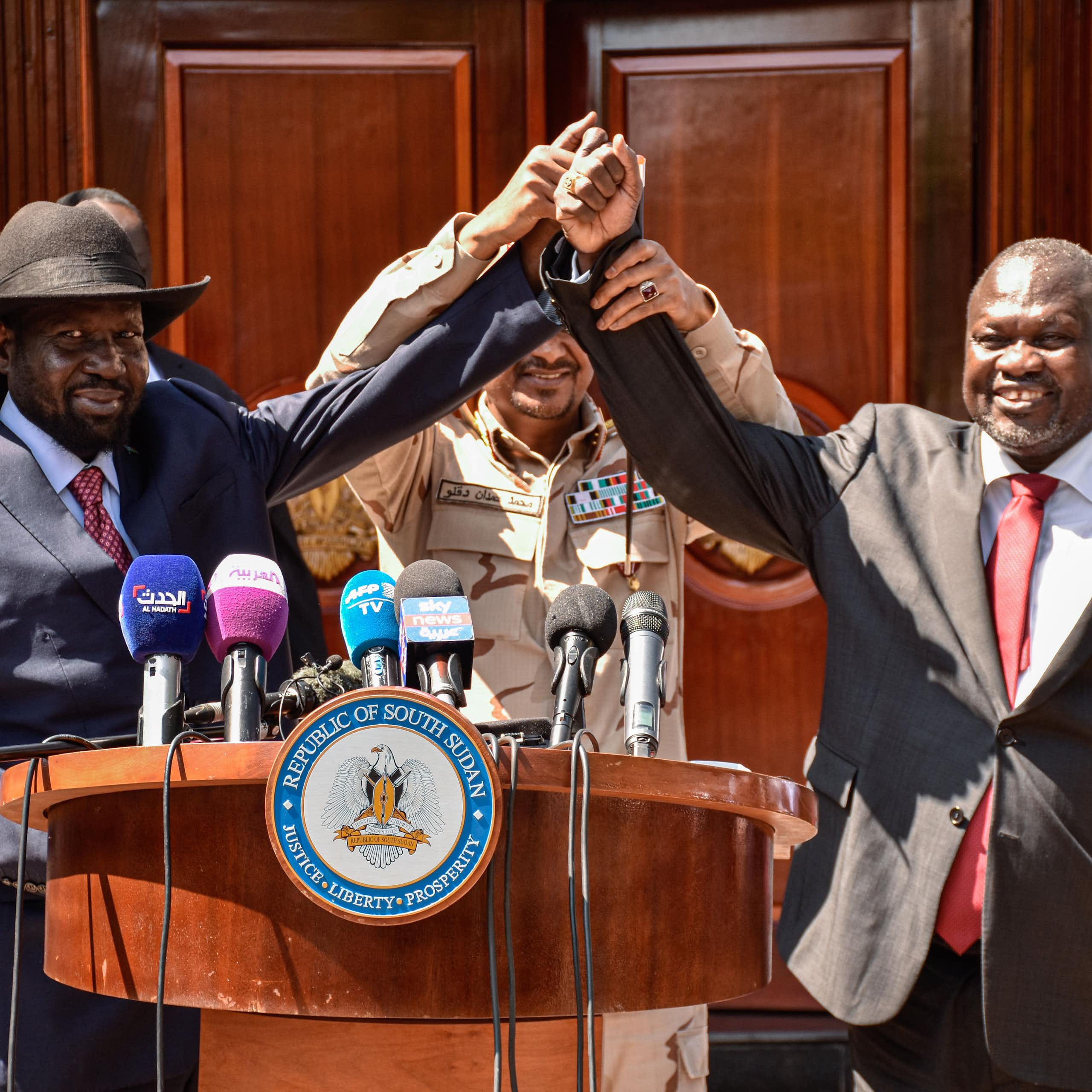 Two men have their hands held together in the middle by a third man as they stand behind a podium with microphones on it