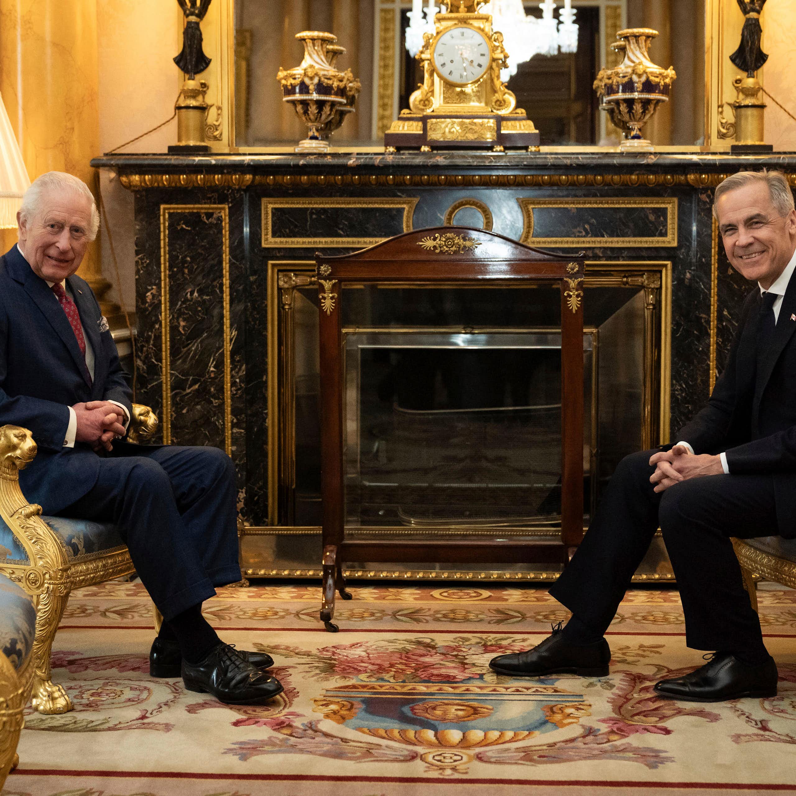 An older man with grey hair sits in an ornate room across from a younger man, also with grey hair. Both smile at the camera.