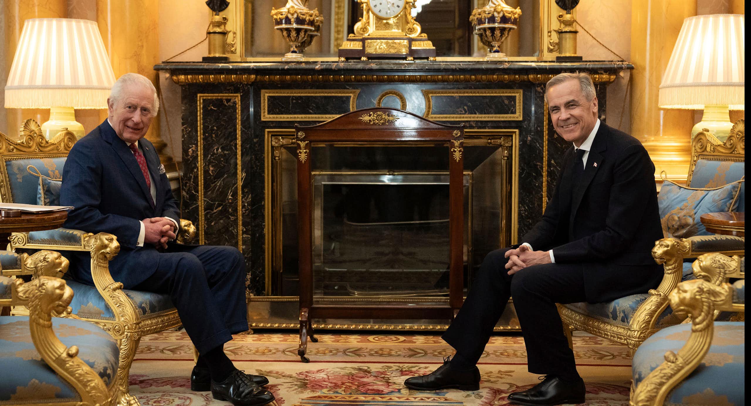 An older man with grey hair sits in an ornate room across from a younger man, also with grey hair. Both smile at the camera.