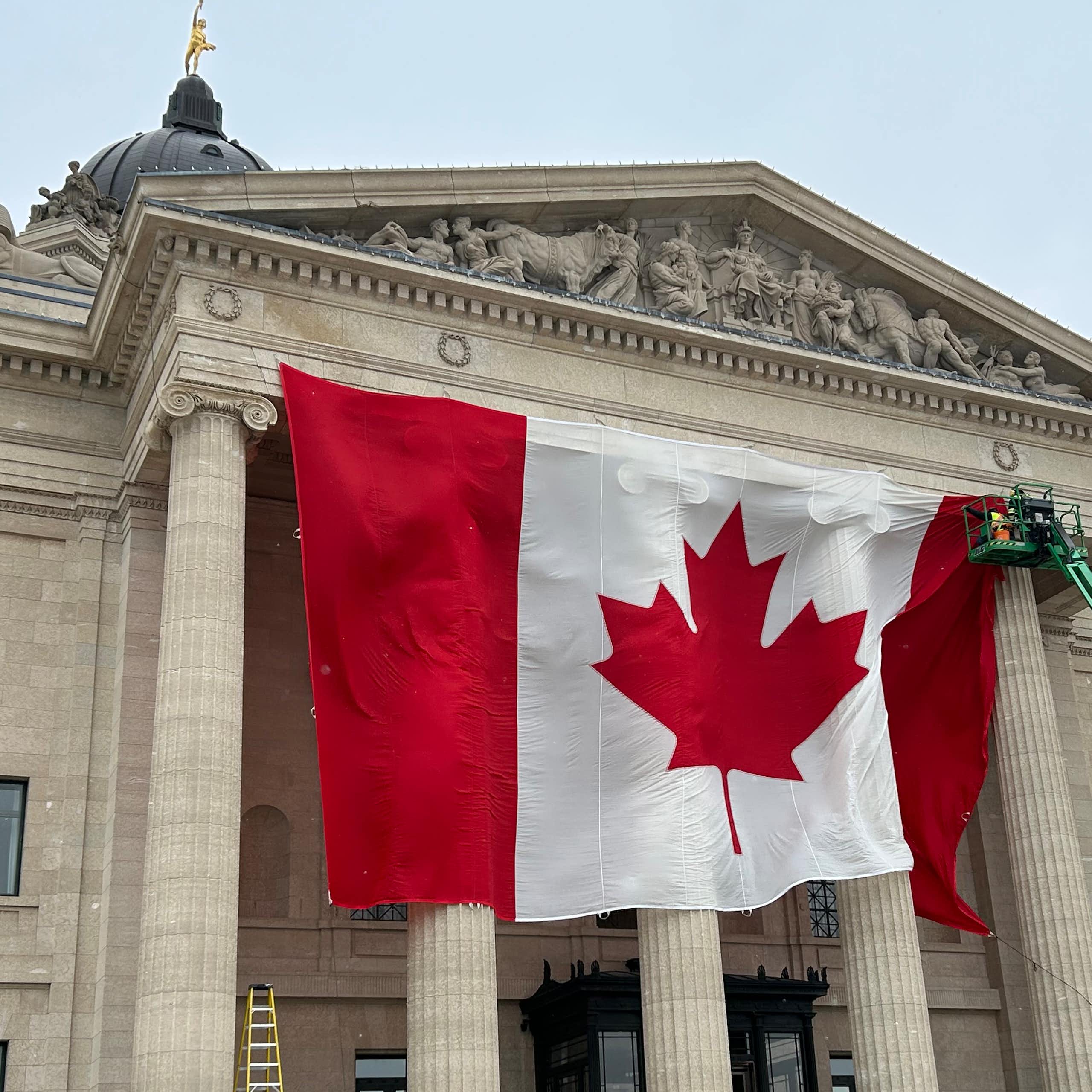 A large Canadian flag is seen being mounted by a crane in front of a tall stone building with columns.