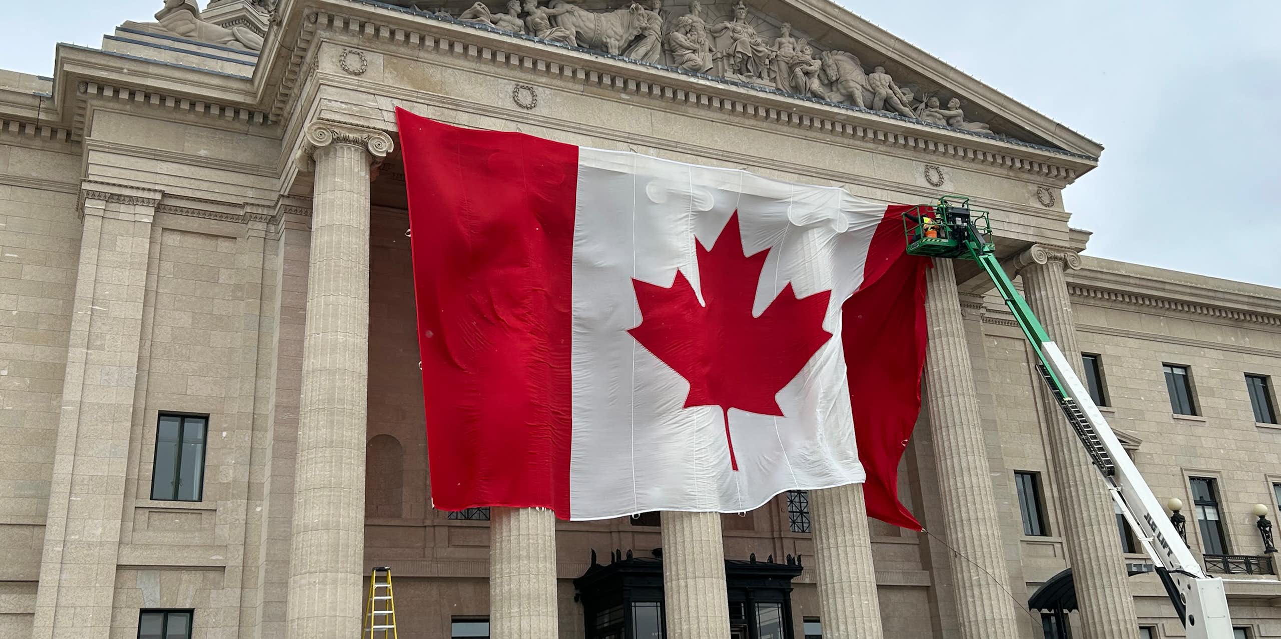 A large Canadian flag is seen being mounted by a crane in front of a tall stone building with columns.