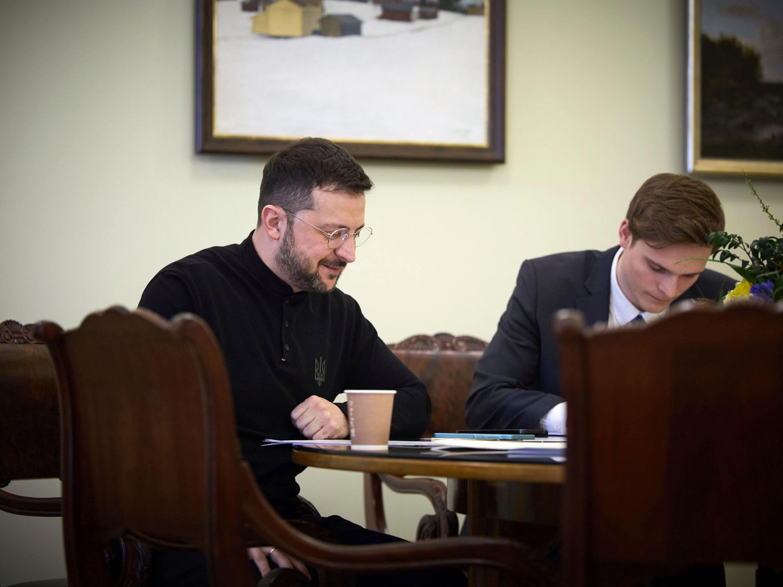 Ukrainian president, Volodymyr Zelensky, sits at a table talking on the telephone while an aide listens in.