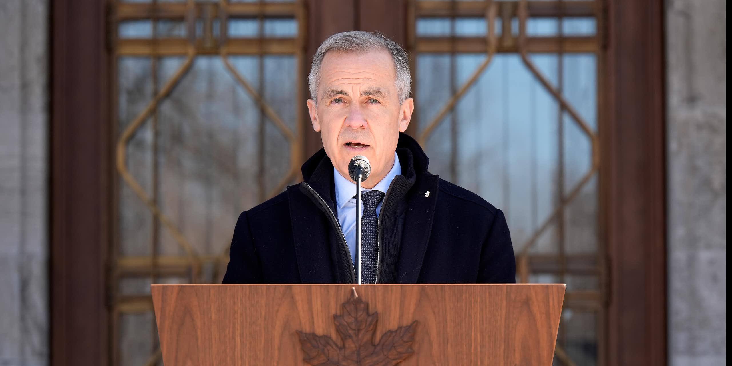 A man with short grey hair speaks into a microphone behind a wooden lectern with a carved maple leaf.