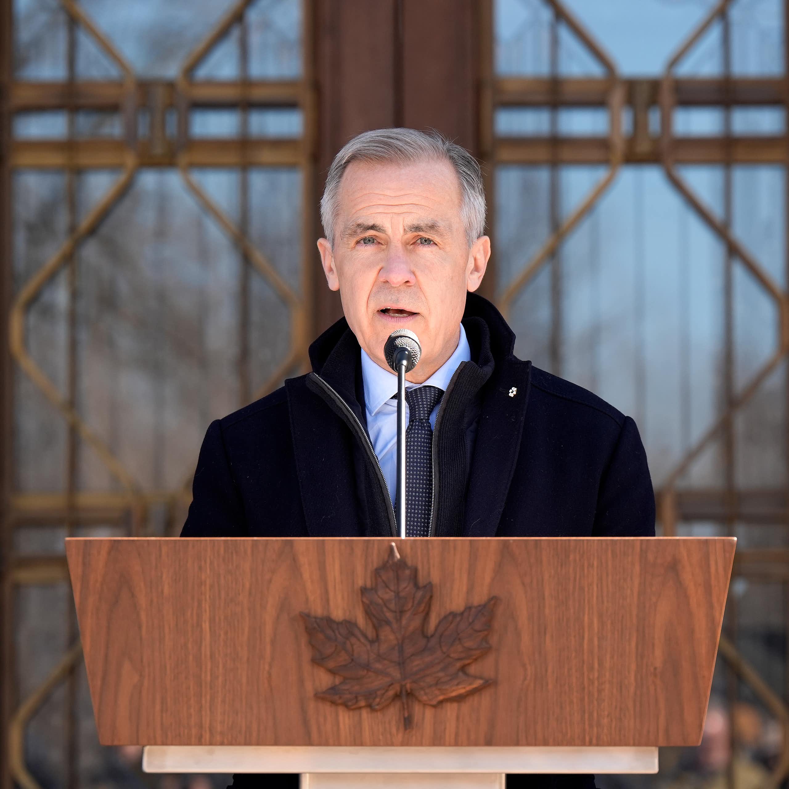 A man with short grey hair speaks into a microphone behind a wooden lectern with a carved maple leaf.