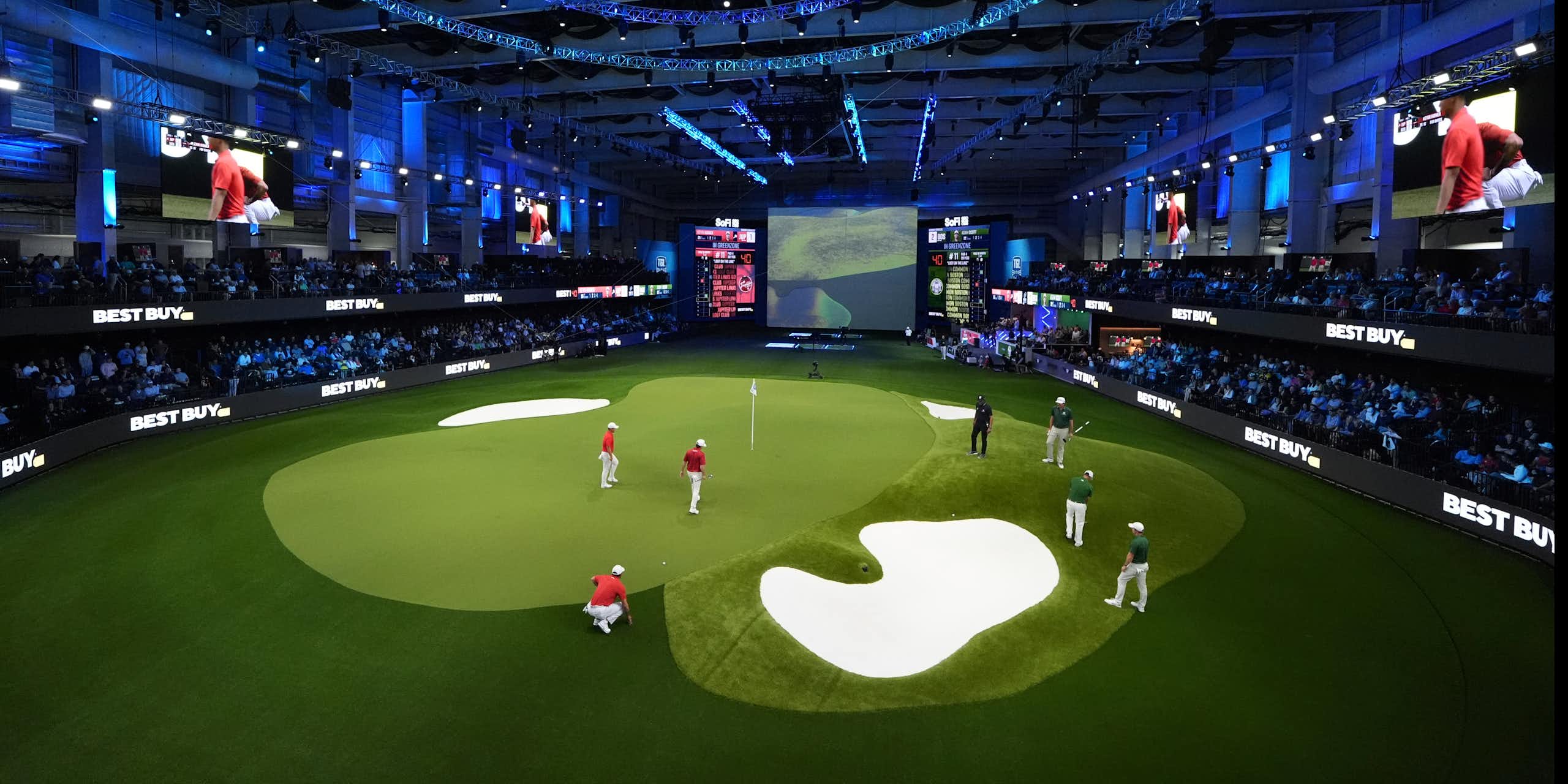 People on an indoor golf green surrounded by stands with an audience