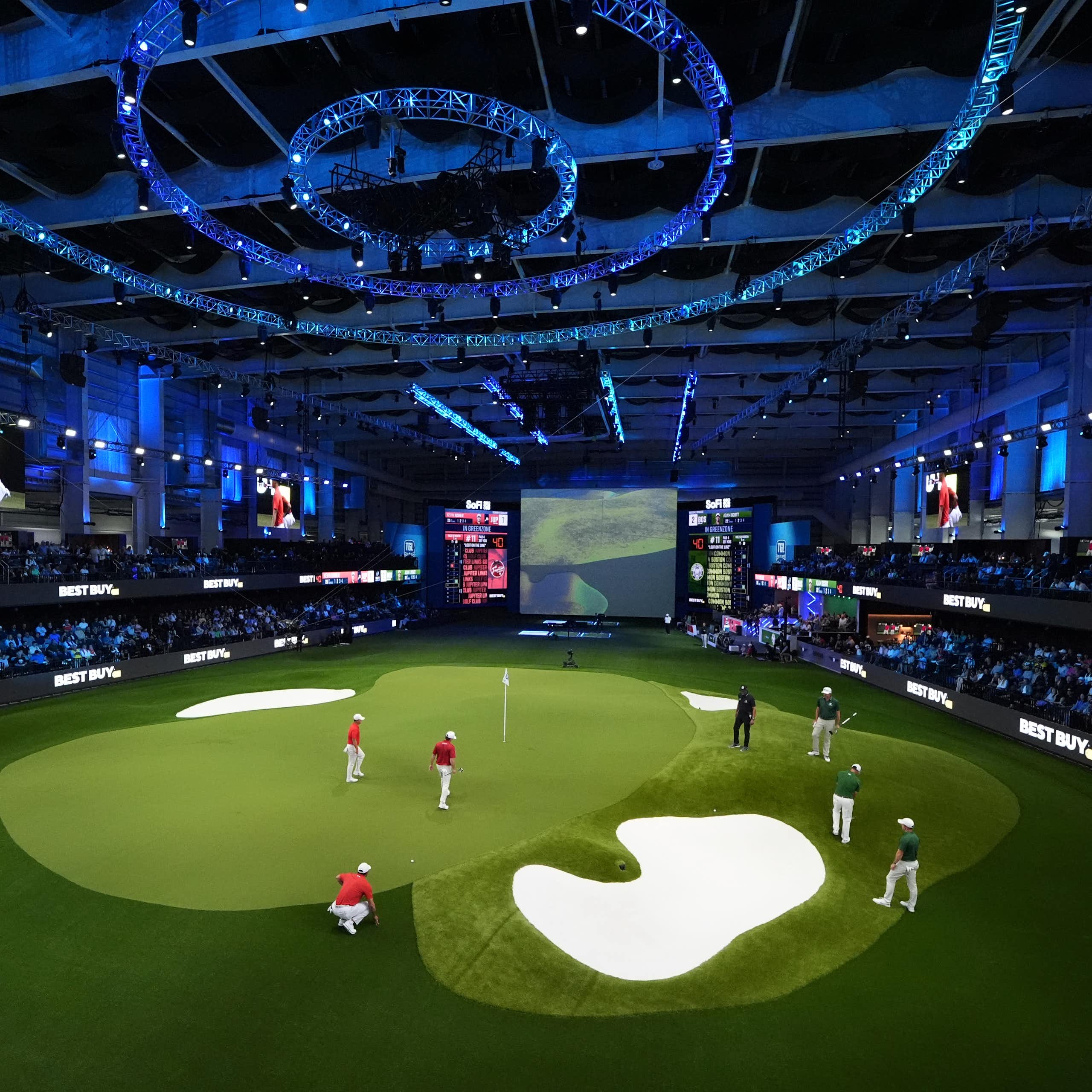 People on an indoor golf green surrounded by stands with an audience