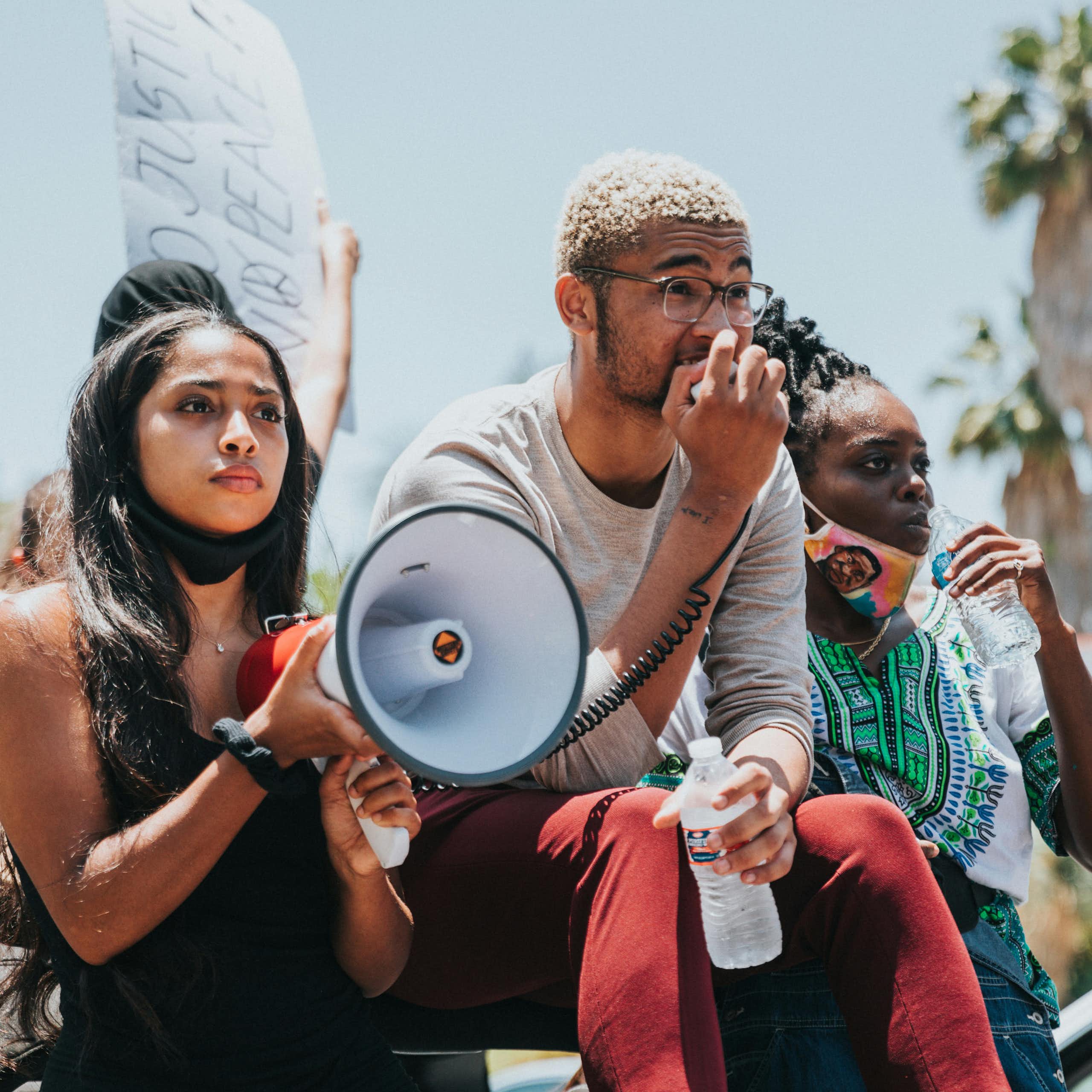 Three people at a demonstration. One person is holding a megaphone while another speaks into the microphone. Another person in the frame is taking a sip of water. Two of the people have masks on, but not covering their faces. 