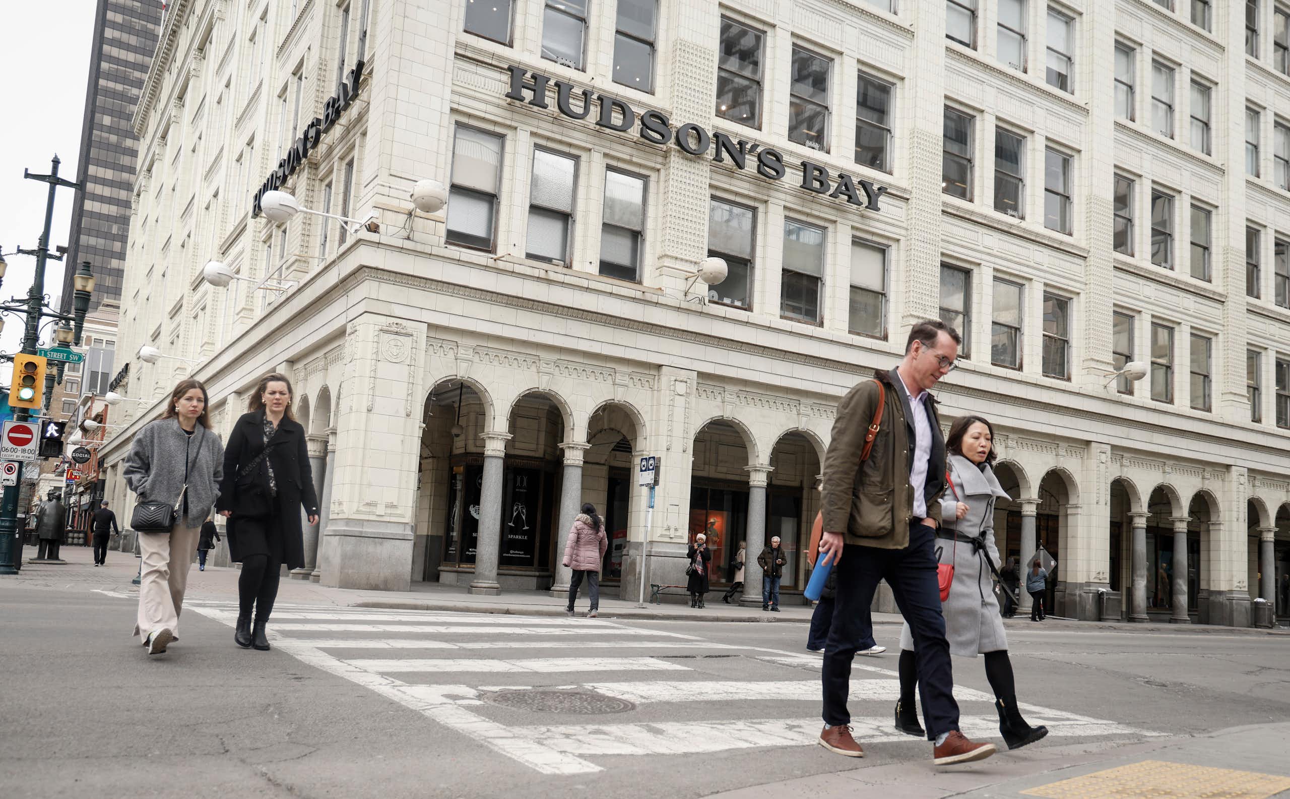 People walk across a city street with a white stone building behind them that reads Hudson's Bay.