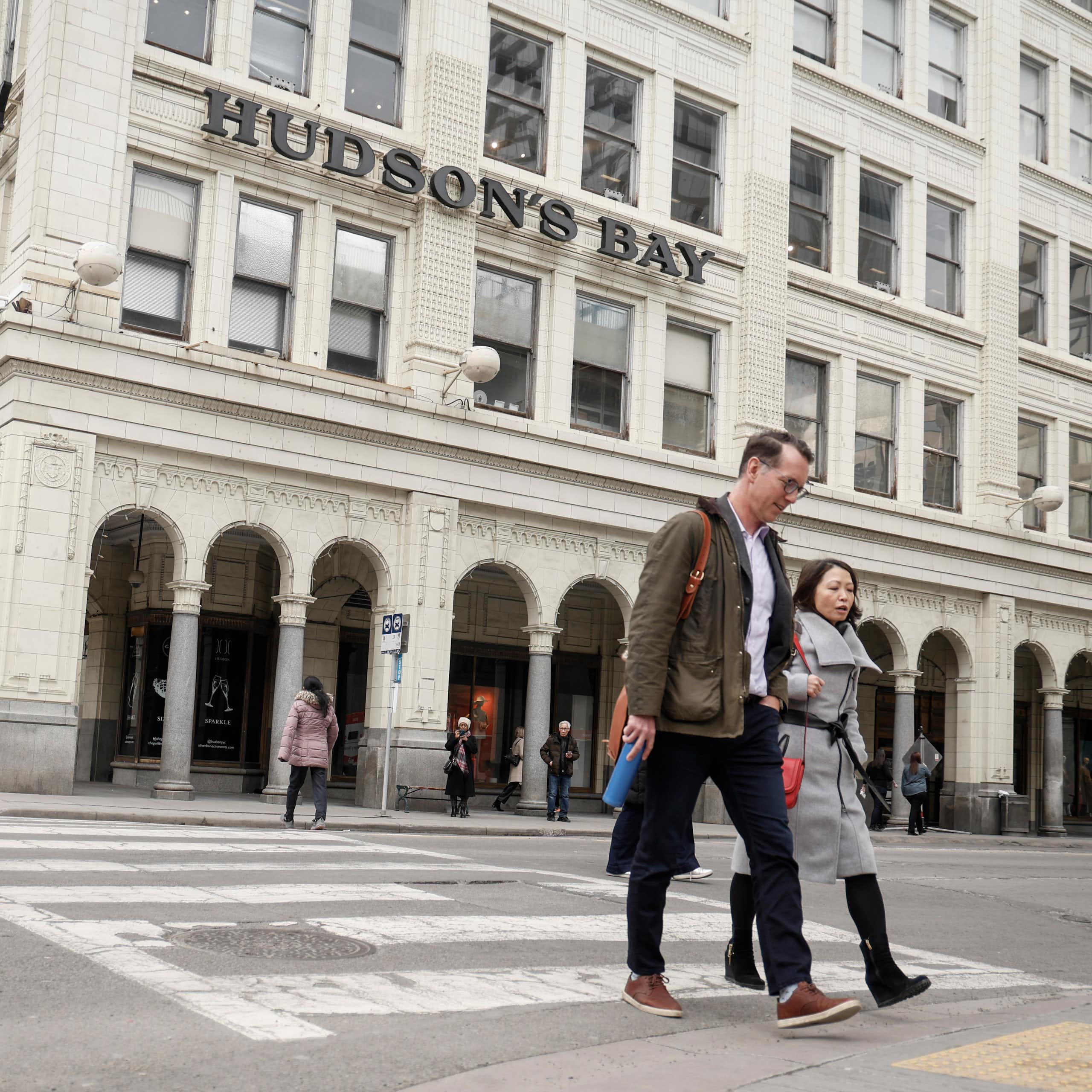 People walk across a city street with a white stone building behind them that reads Hudson's Bay.