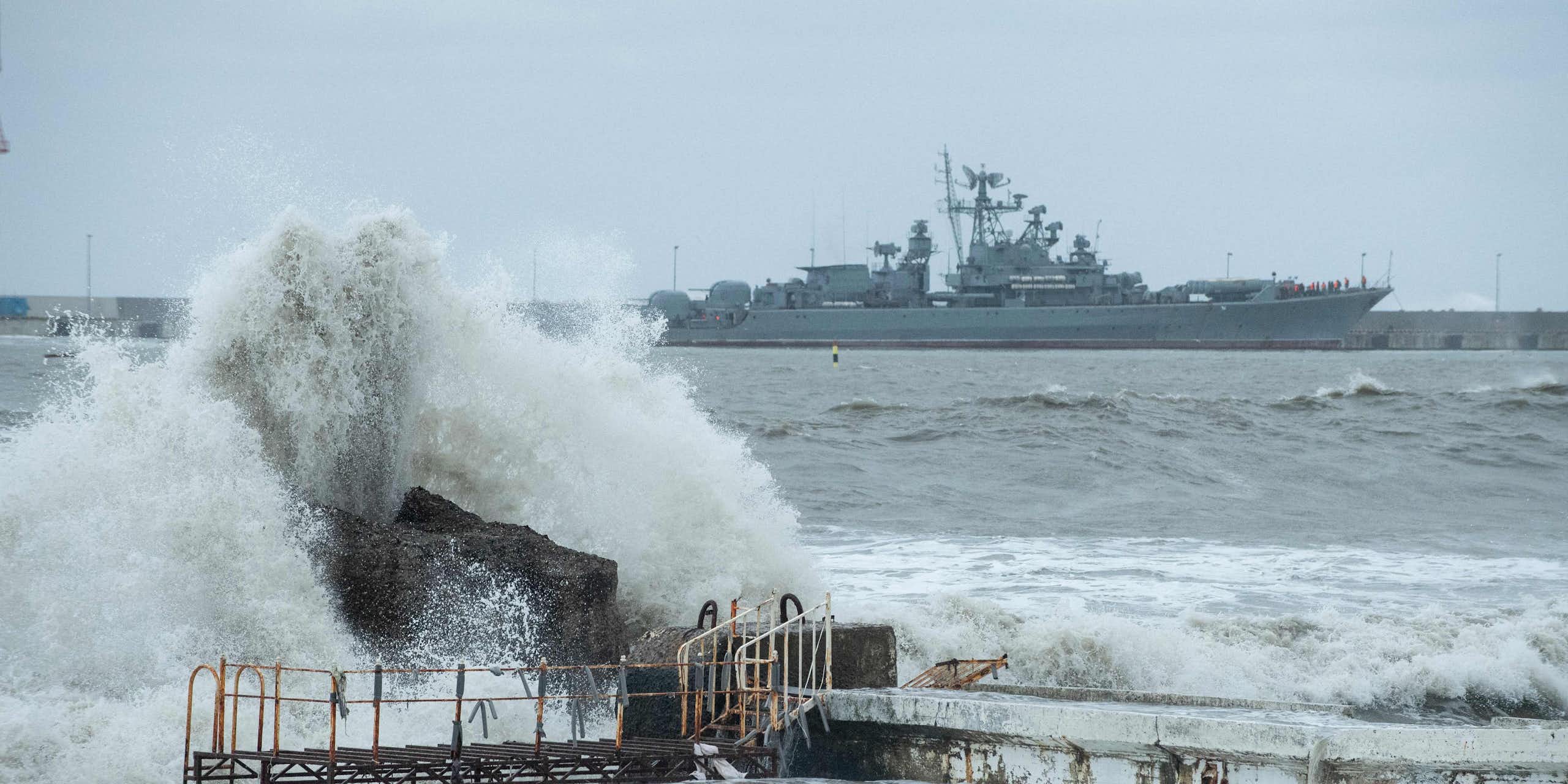 A large warship is seen at sea behind a big wave crashing on some rocks
