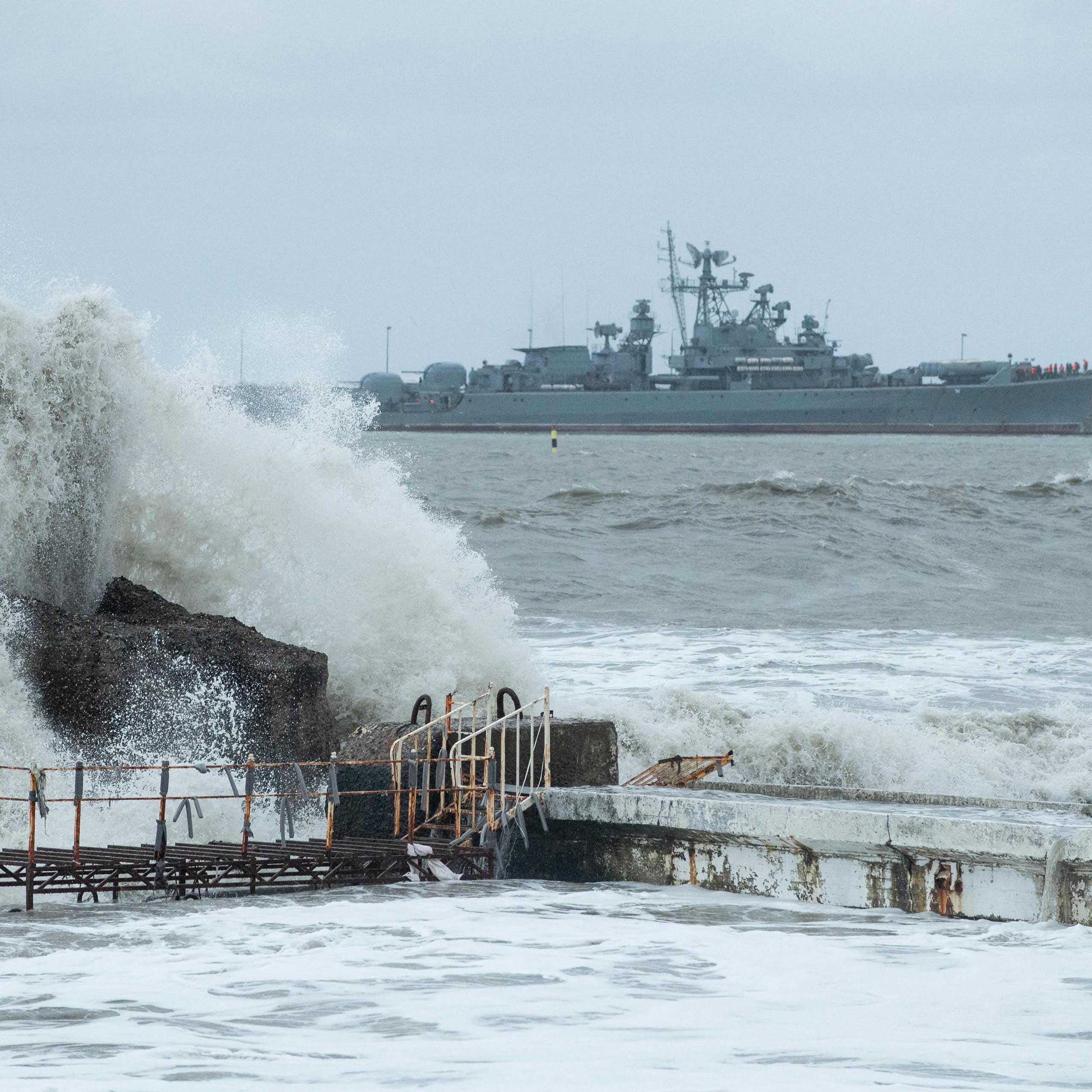 A large warship is seen at sea behind a big wave crashing on some rocks