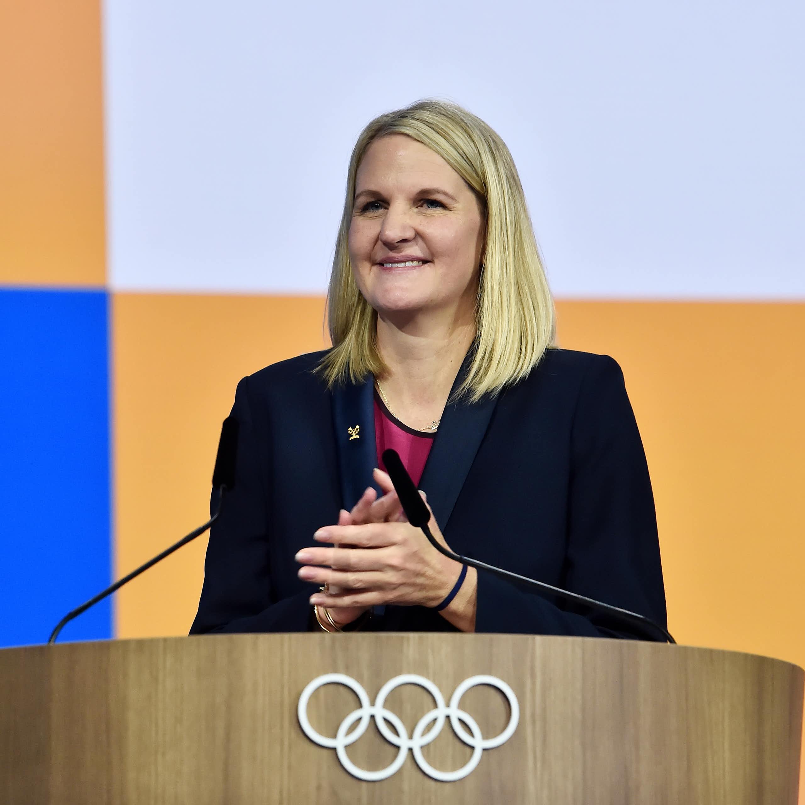 A woman with long blonde hair stands at a podium decorated with the Olympic logo of five rings. She's smiling.