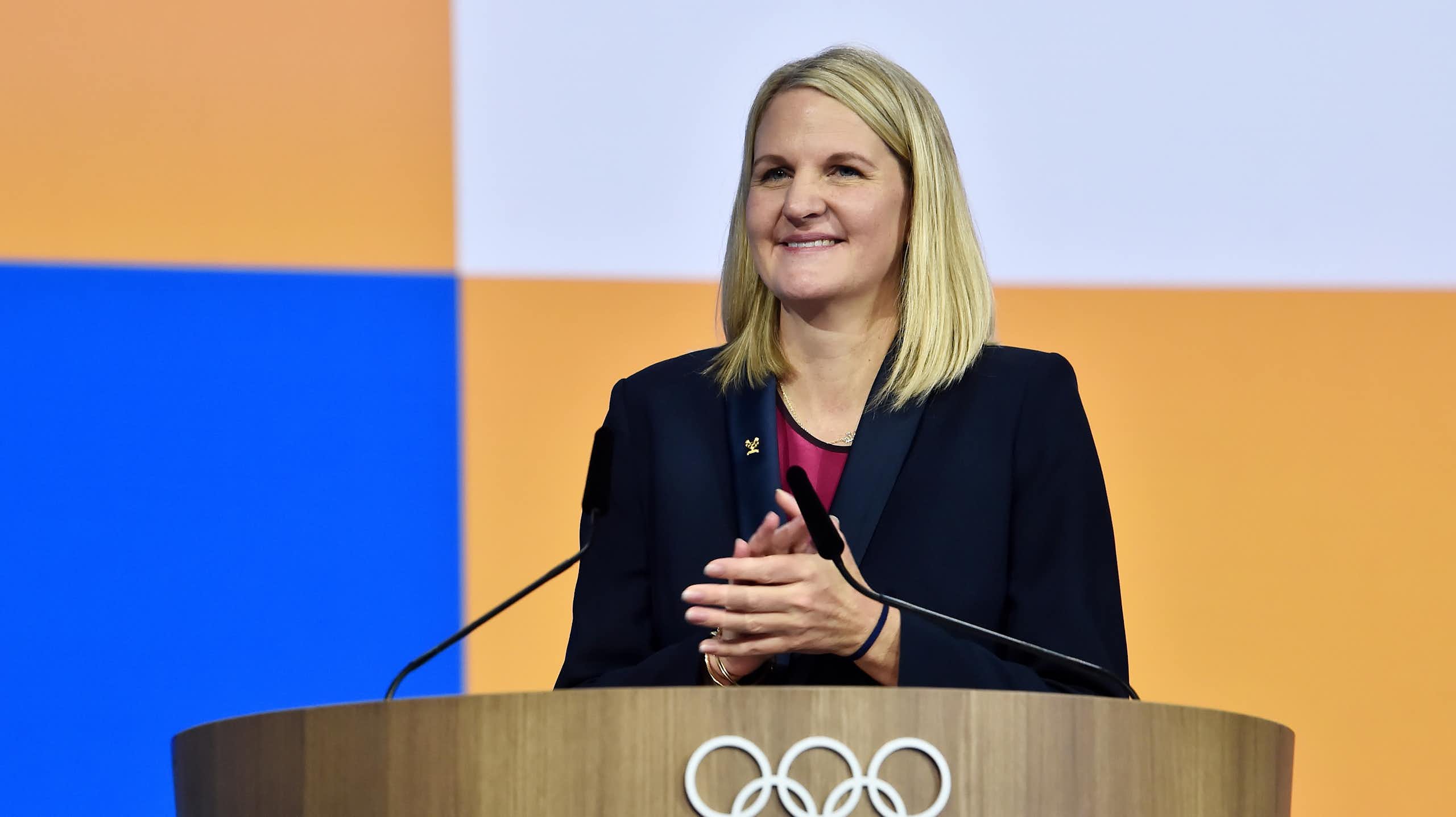 A woman with long blonde hair stands at a podium decorated with the Olympic logo of five rings. She's smiling.