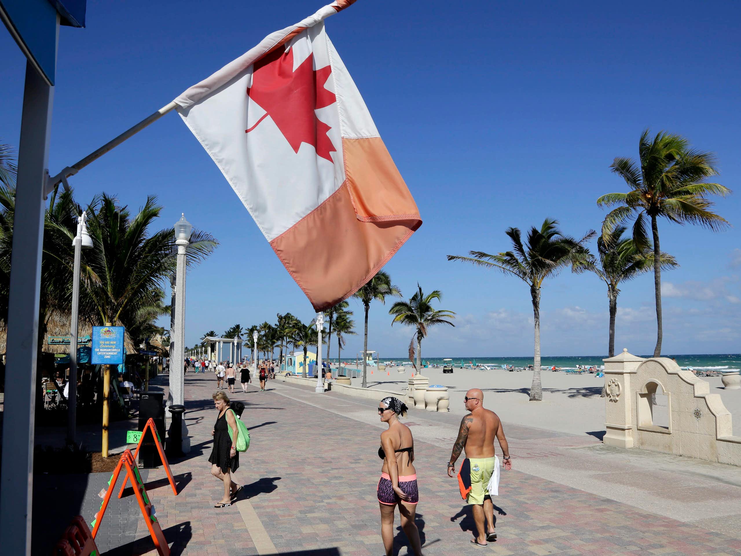 Un drapeau canadien flotte près d'une plage de sable, tandis que des gens marchent le long d'une promenade.