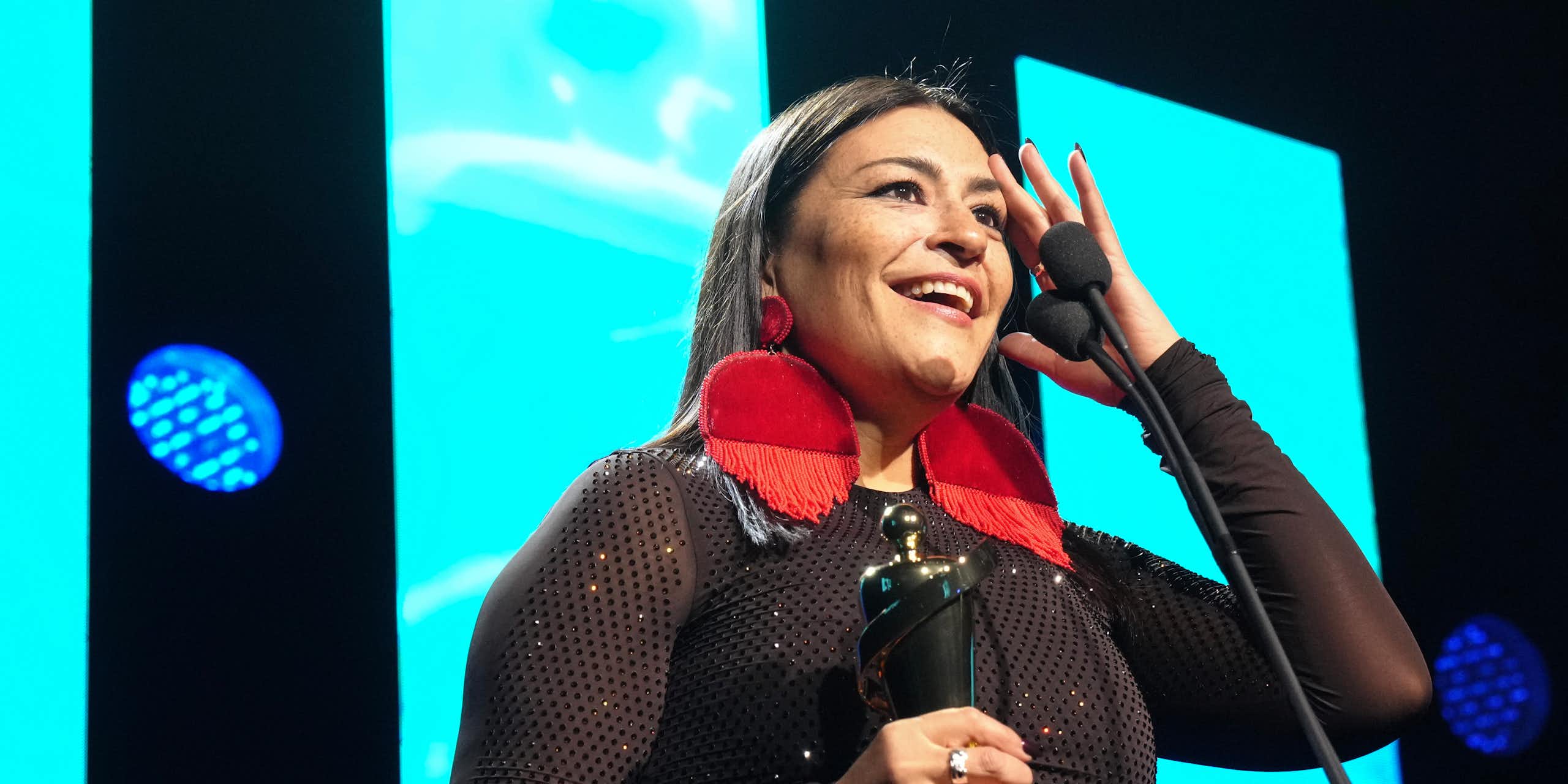 A woman with circular red earrings at microphone holding trophy.