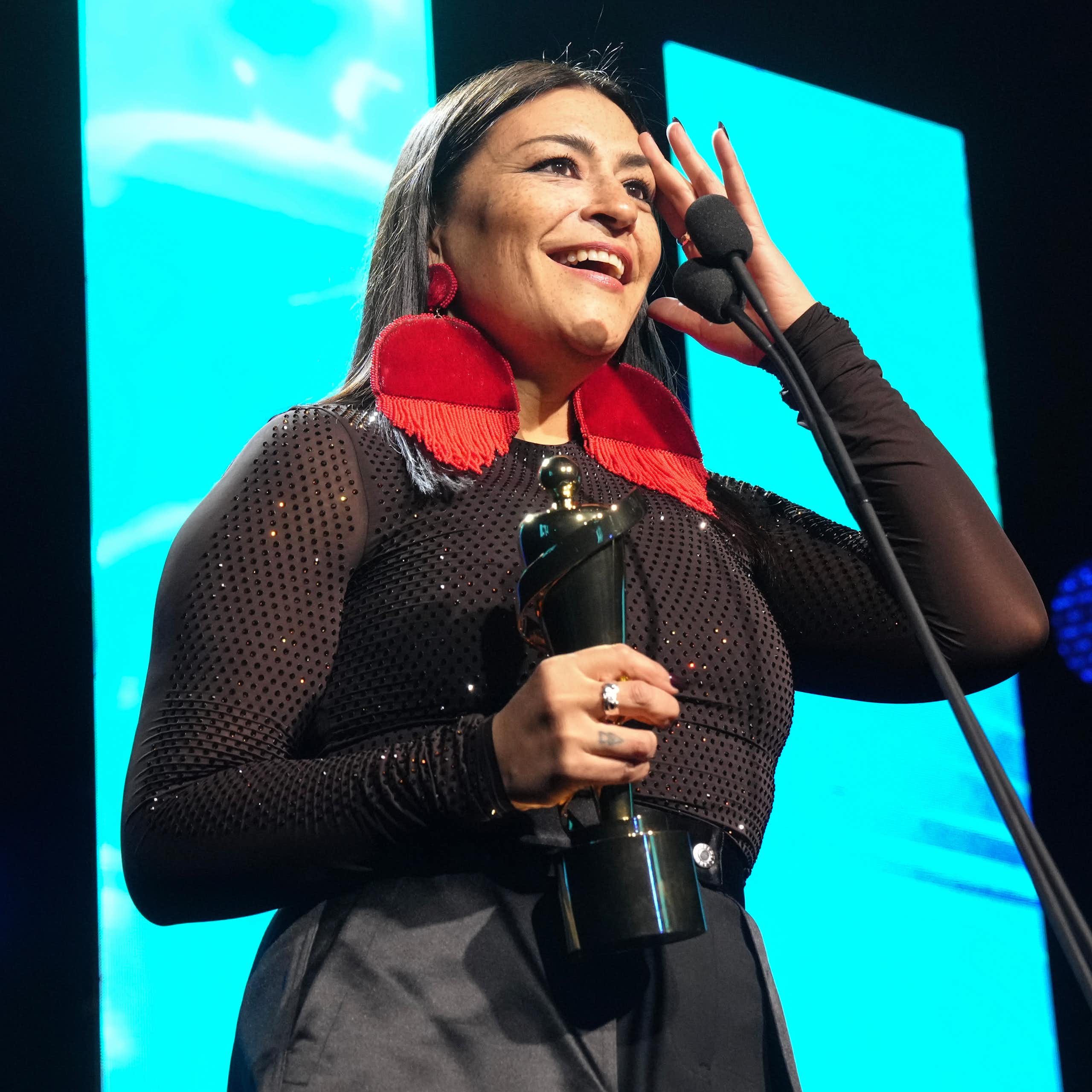 A woman with circular red earrings at microphone holding trophy.