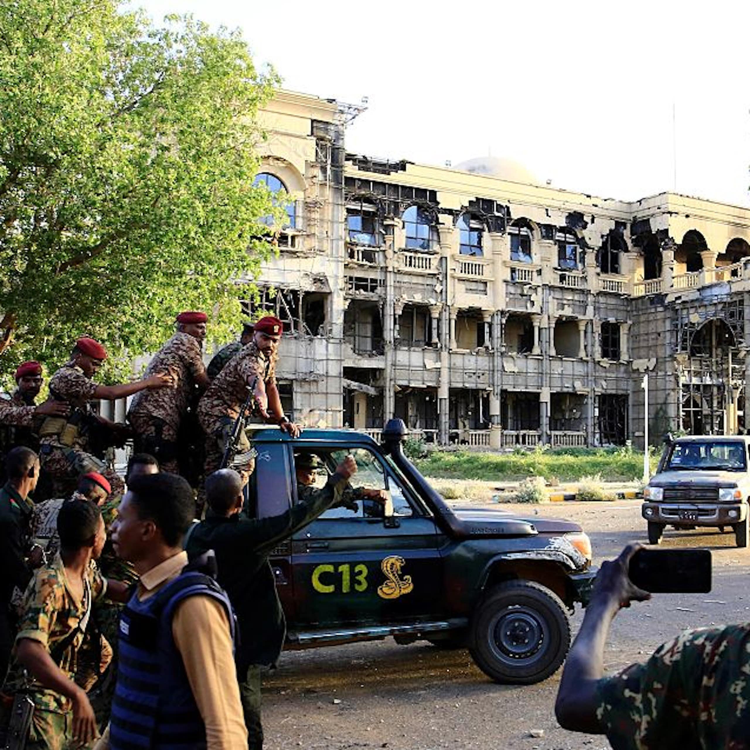 Men in military fatigues aboard a pickup vehicle written C13 drive near a three-storey building that has a damaged exterior.